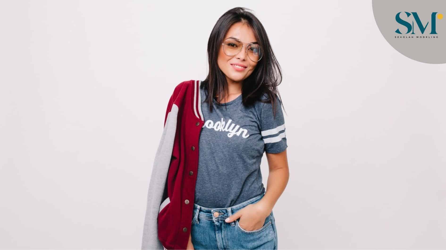 Slim joyful girl in gray t-shirt and denim pants posing with hand in pocket and smiling. Black-haired female model in jeans and bomber standing