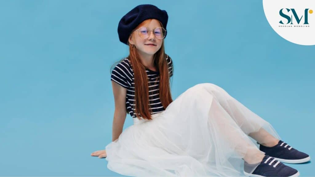 Cool young parisian in good mood in eyeglasses long white skirt and dark sneakers sitting on floor and posing on blue backdrop